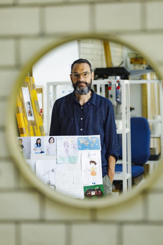 A photograph of Sam Durant in the reflection of a round mirror in his studio. Sam has dark hair, a moustache and beard, and glasses.
