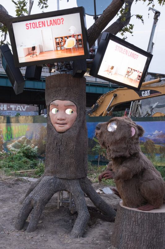 A sculture of a tree trunk with a human face stands next to a beaver sculpture; above them, screens display the question “STOLEN LAND?” over a photo of a modern interior.