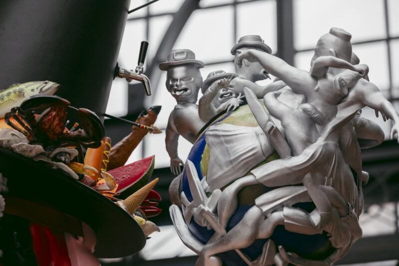 A sculptural artwork features silver figures in hats atop a globe, surrounded by various colorful objects.