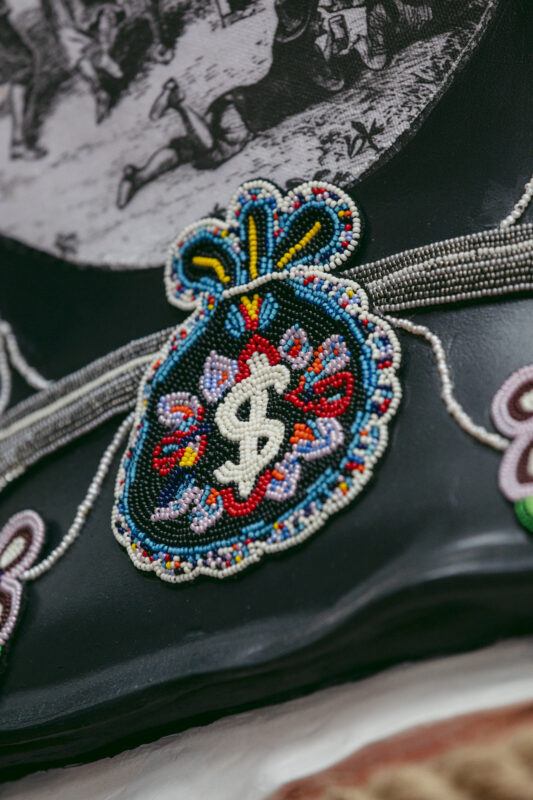 Beaded patch in the shape of a money bag with a dollar sign, sewn onto a dark fabric background with decorative patterns.