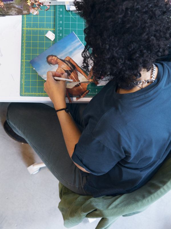Joiri Minaya leaning over a table with a cutting board, holding an exacto knife to cut out an image of a woman in a bathing suit from a magazine.