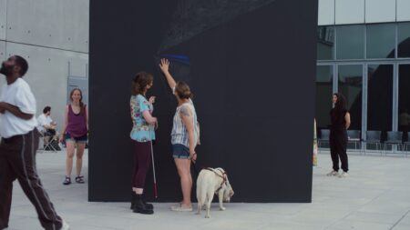 People explore a large black wall on an outdoor pavilion; two women stand in front of the wall, one touches it while holding a guide dog, the other uses a white cane. Two other people are nearby, one looking up at the wall.
