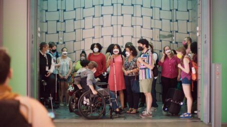A diverse group of people, some wearing masks and one using a wheelchair, stand together in a large elevator with a geometric background.
