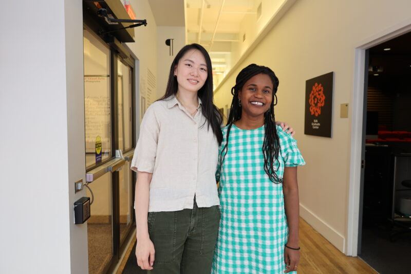 Two women stand side by side in a hallway, smiling at the camera. One wears a beige shirt and green pants, the other a green checkered dress.
