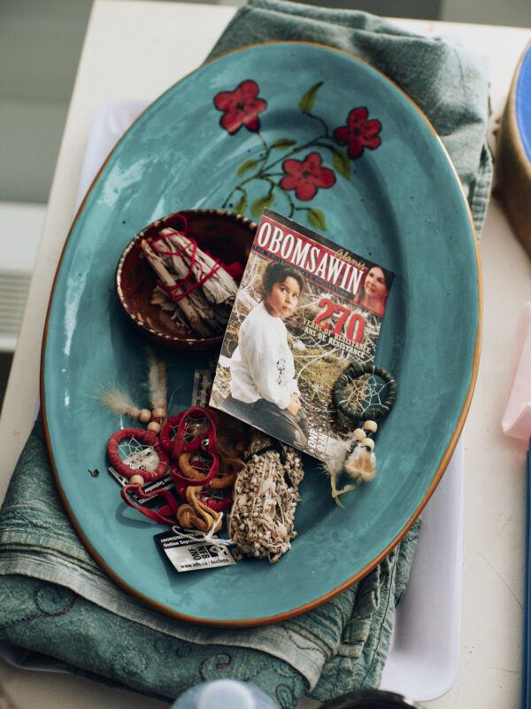 A turquoise platter with a floral design holds a magazine featuring Alanis Obomsawin, small bowls with herbs, dreamcatchers, feathers, and other decorative objects.