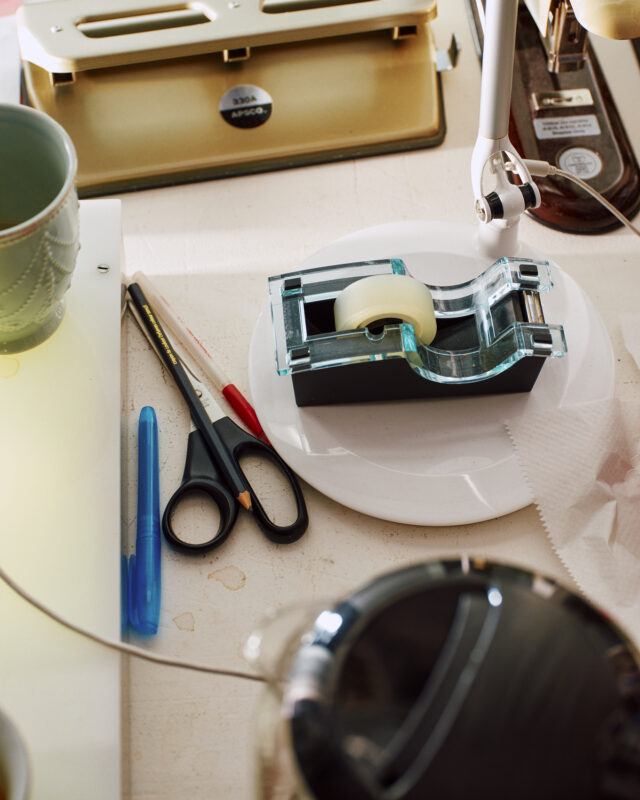 A desk with scissors, a blue pen, a pencil, a tape dispenser on a plate, a green cup, a lamp, and a beige hole punch.