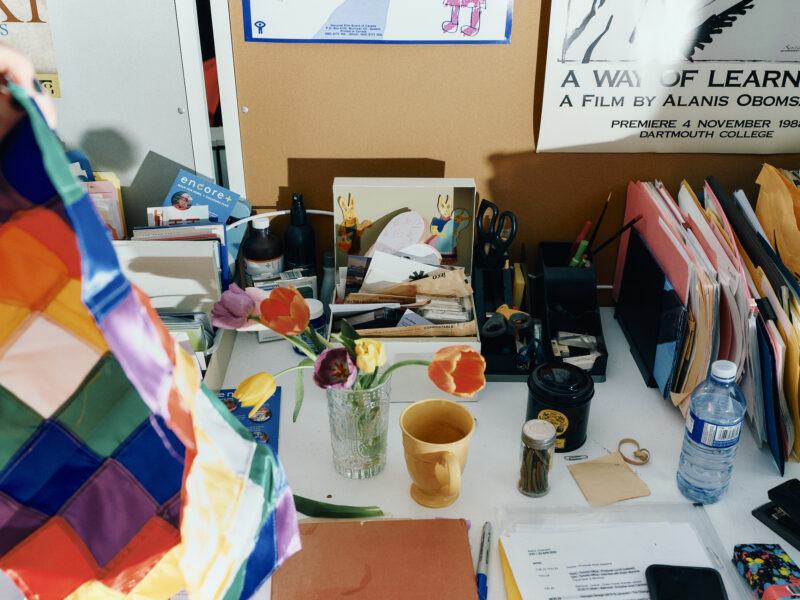 A cluttered desk with papers, folders, a vase of tulips, a coffee cup, a water bottle, office supplies, and posters and notes pinned to the wall behind it.
