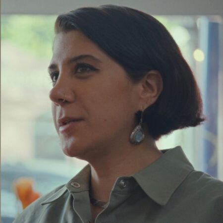Morehshin Allahyari with short dark hair and earrings is wearing a collared shirt, looking slightly to the side indoors with a blurred background.