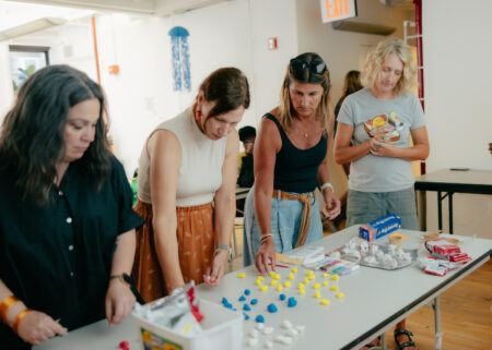 Four women stand at a table organizing small yellow and blue objects, with craft supplies and cartons on the table in a well-lit room.