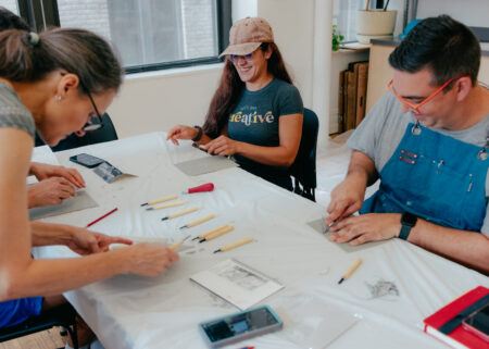 Three adults sit at a table using carving tools on small pieces of linoleum.