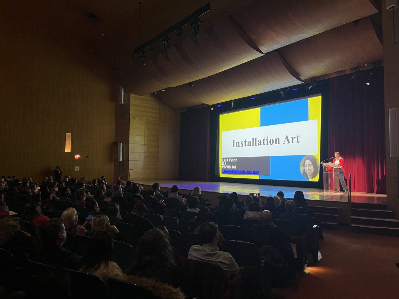 A speaker presents a slideshow titled "Installation Art" to an audience in a large auditorium with wooden walls and ceiling.