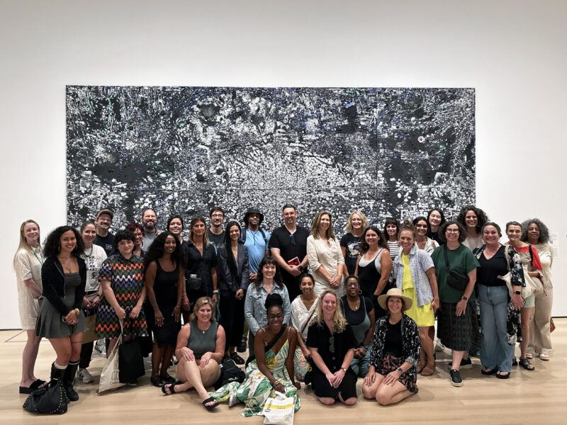 A group of people pose together in front of a large abstract black and white artwork in a gallery setting.