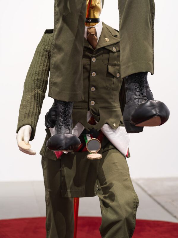 A mannequin in an olive green military uniform, with another mannequin's legs and boots suspended in front, positioned on a red circular platform.