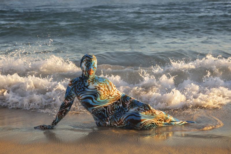 A person in a patterned bodysuit is lying on a sandy beach with waves approaching.
