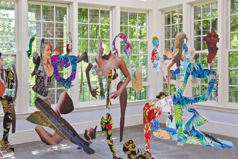 Hanging cut outs of abstract human figures with floral patterns suspended in a sunlit room with large windows.