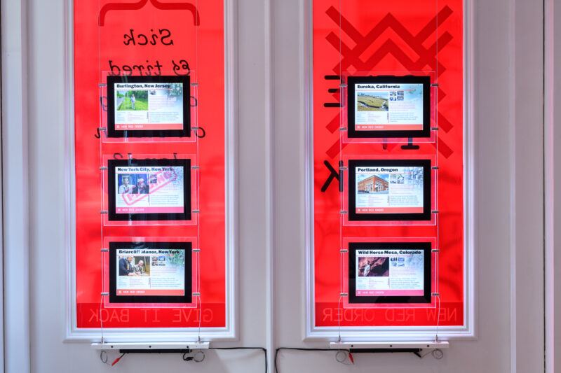 Six screens are mounted on a bright red wall, each displaying information about different U.S. cities or areas, including photos and text.