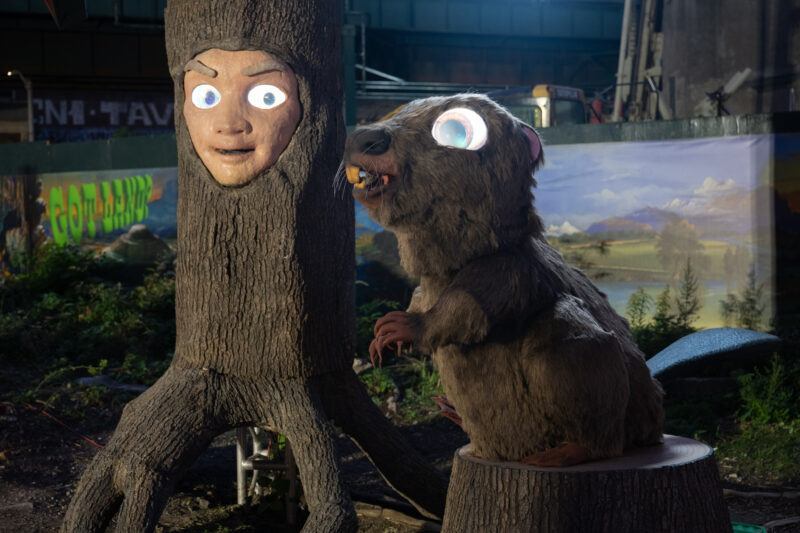 A scultprue of a tree trunk with a human face and glowing eyes stands next to a beaver with illuminated eyes in an outdoor setting with artificial landscaping.