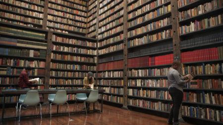 Three people read books in a large library filled with tall, packed bookshelves and rows of chairs and tables on a wooden floor.