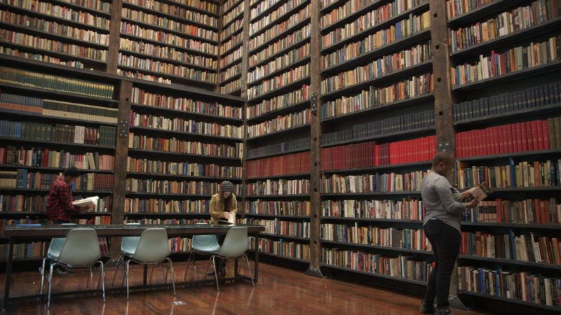 Three people read books in a large library filled with tall, packed bookshelves and rows of chairs and tables on a wooden floor.