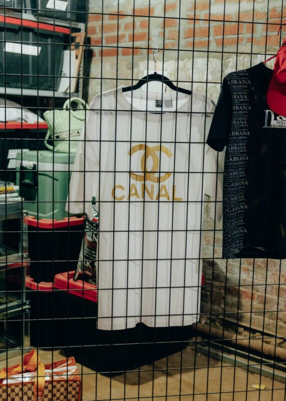 A white t-shirt hanging off of a metal grate fence, with a double-c logo and CANAL on it.