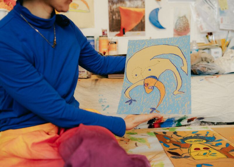 Close up of Tamar Ettun's hands holding up a drawing of a figure twisiting on a blue background.