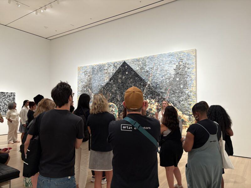 A group of people stands in an art gallery facing a large, abstract artwork with a prominent black geometric shape on the wall.