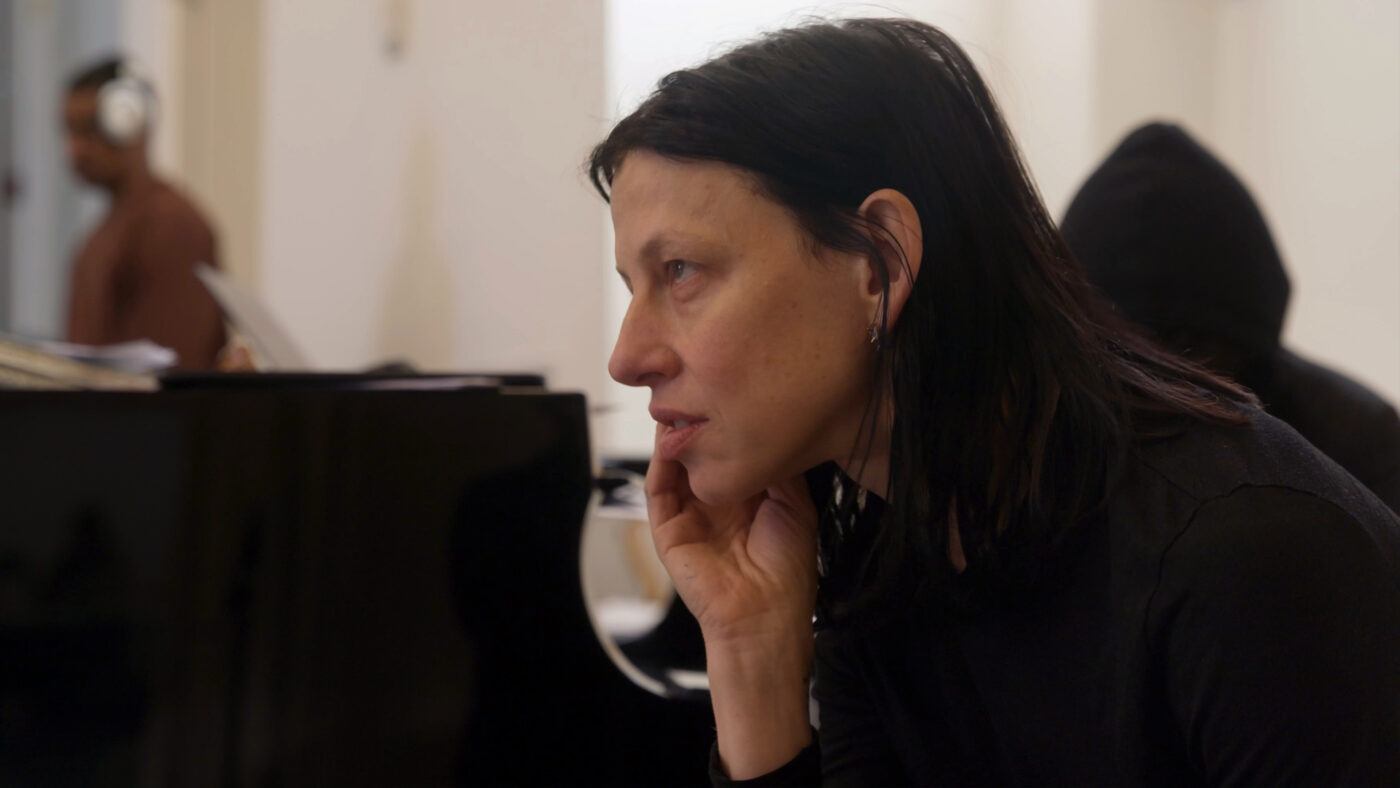 Anne Imhof holding her head in her hand looking forward, with a piano next to her.