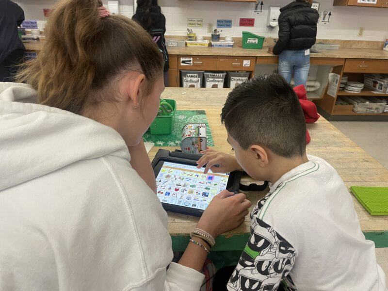 Two students sit at a table using a tablet with picture-based communication software in a classroom setting.