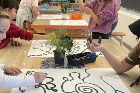 Students sit and stand around a table, drawing abstract black lines on large white papers with markers; potted plants and art supplies are on the table.