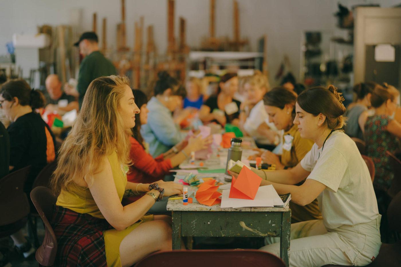 A long table of Art21 Educators slightly out of focus, with two educators on the end of the table in focus. On the table in front of them is bookmaking supplies.