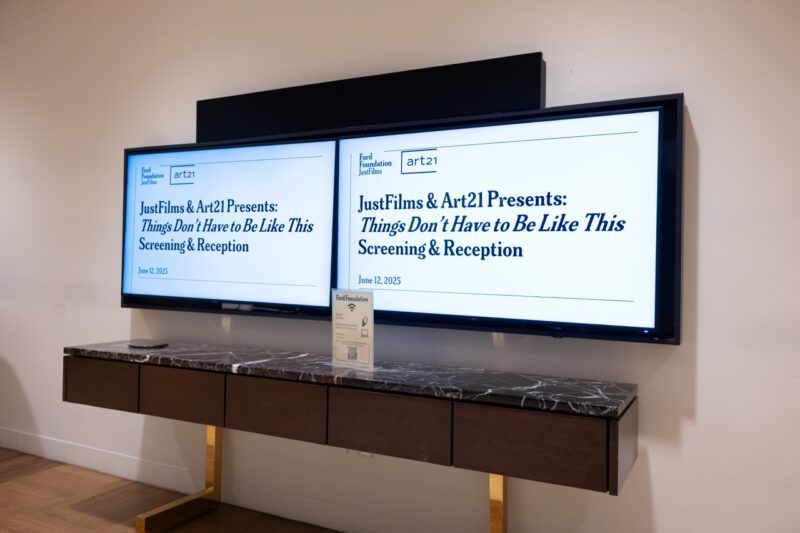 Two large monitors display event information for "Things Don’t Have to Be Like This Screening & Reception" by JustFilms & Art21, mounted above a marble-topped console table.