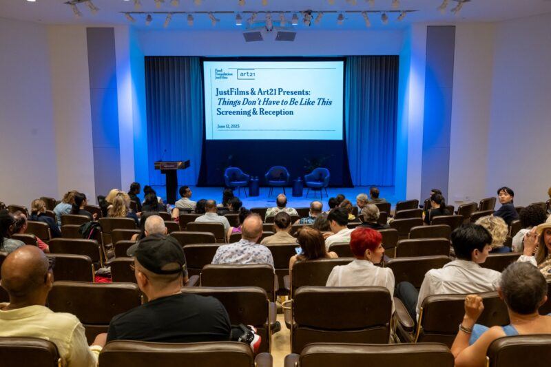 Audience seated in an auditorium faces a stage with two chairs and a screen displaying event details for a screening and reception titled “Things Don’t Have to Be Like This.”.