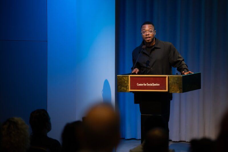 A man speaks at a podium labeled "Center for Social Justice" in front of an audience, with blue lighting in the background.