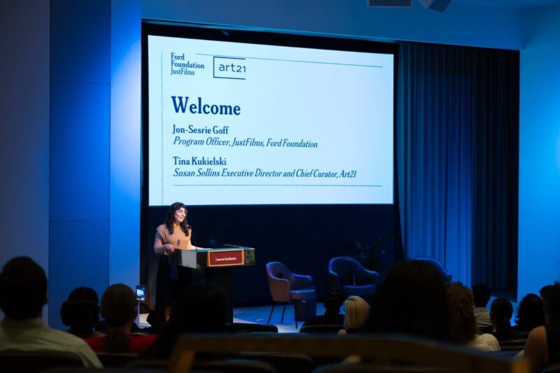 A woman stands at a podium addressing an audience in an auditorium; a large screen behind her displays a welcome message for a Ford Foundation and Art21 event.