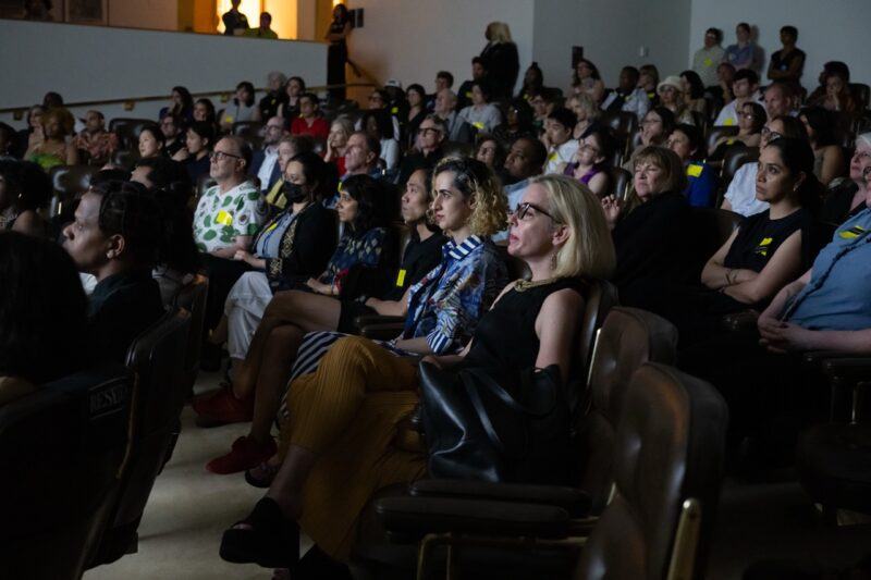 A diverse audience sits in a dimly lit auditorium, attentively watching something on stage or screen.