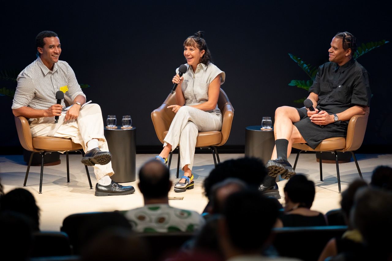 Three people sit on stage in chairs, each holding a microphone and speaking to an audience.