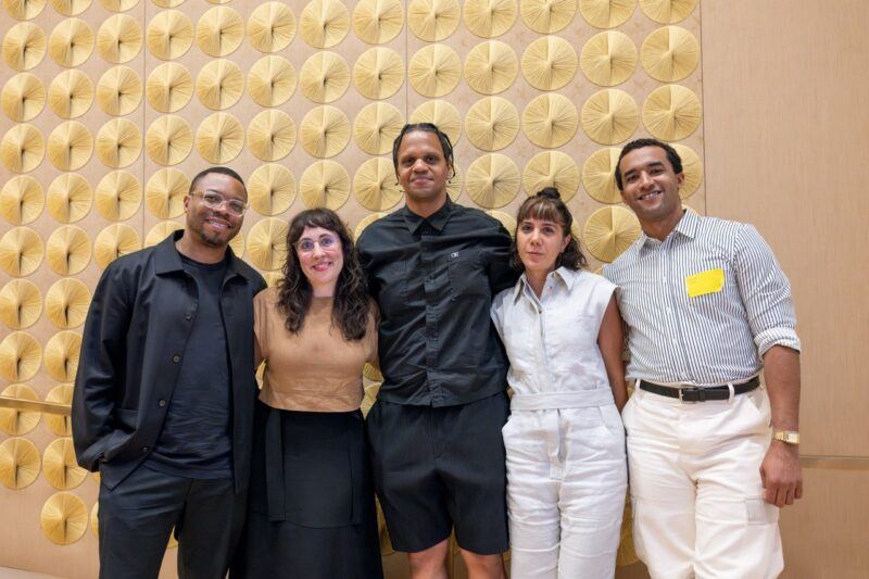 Five people standing together in front of a beige wall decorated with circular patterns, smiling and posing for a group photo.