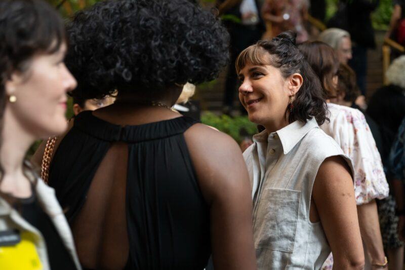 A group of people stand close together at an indoor event, conversing and smiling amid a busy background.