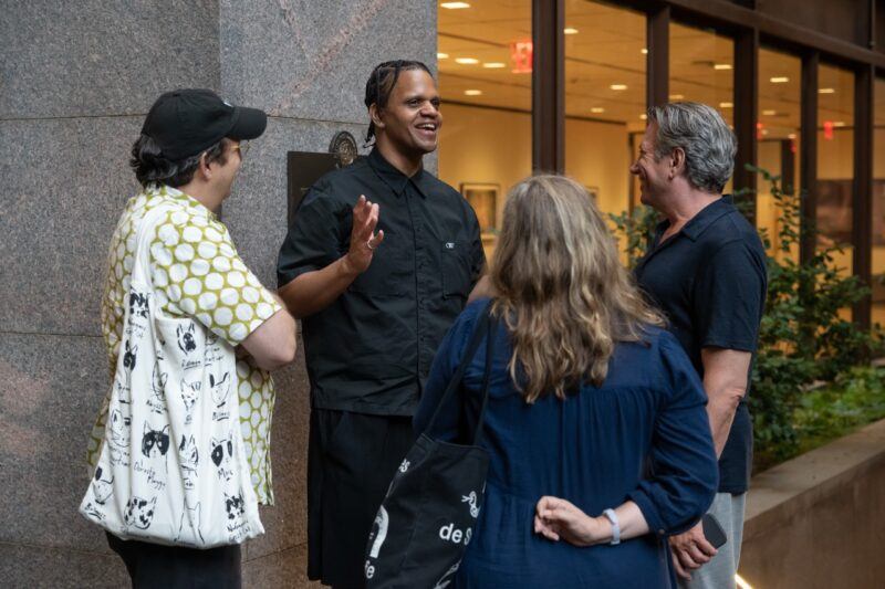 Four people stand in a group outdoors, engaged in conversation near a stone building with large windows and plants in the background.