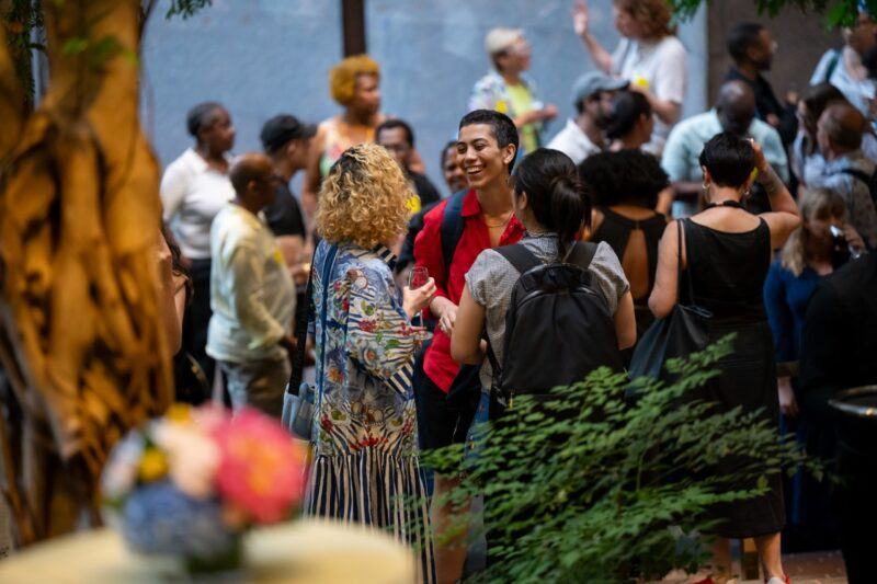 A diverse group of people socializes at an indoor event, with some conversing and others mingling; plants and floral arrangements are visible in the foreground.