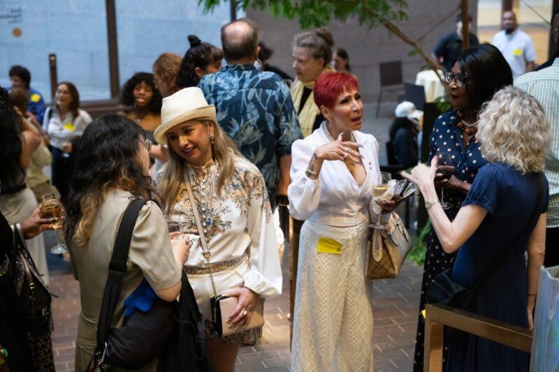 A group of people stand and converse at an indoor social event, some holding drinks, with seating and greenery visible in the background.