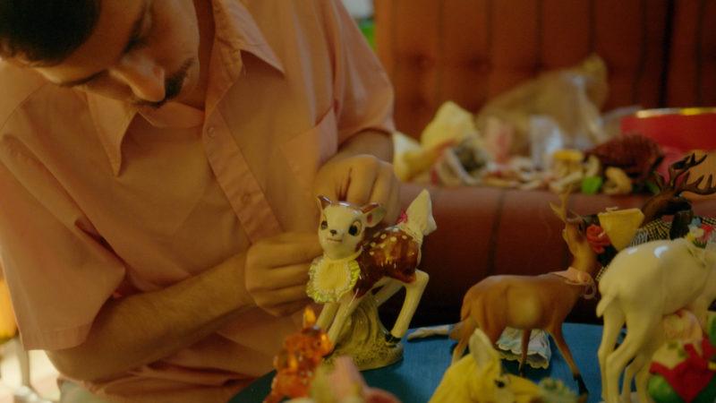 Adam Milner bent over a blue table tying a bib a ceramic deer figurine. There are several other deer figurines on the table.