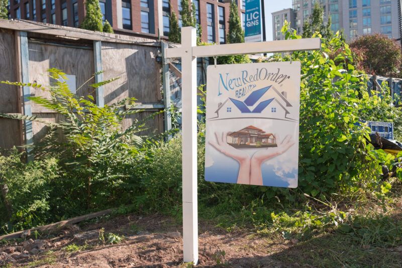 A real estate sign for “New Red Order Realty” stands in a vacant, overgrown lot with buildings visible in the background.