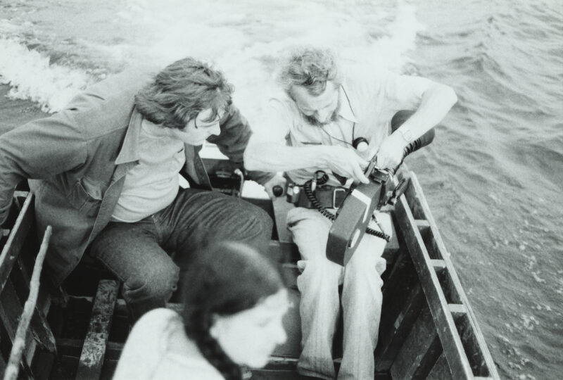 Black and white of photo of two men sitting in a small wooden boat on water, focusing on adjusting a camera, while a woman in braids sits in the foreground.