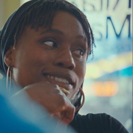 Mimi Onuoha wearing hoop earrings and a headscarf smiles while looking to the side.