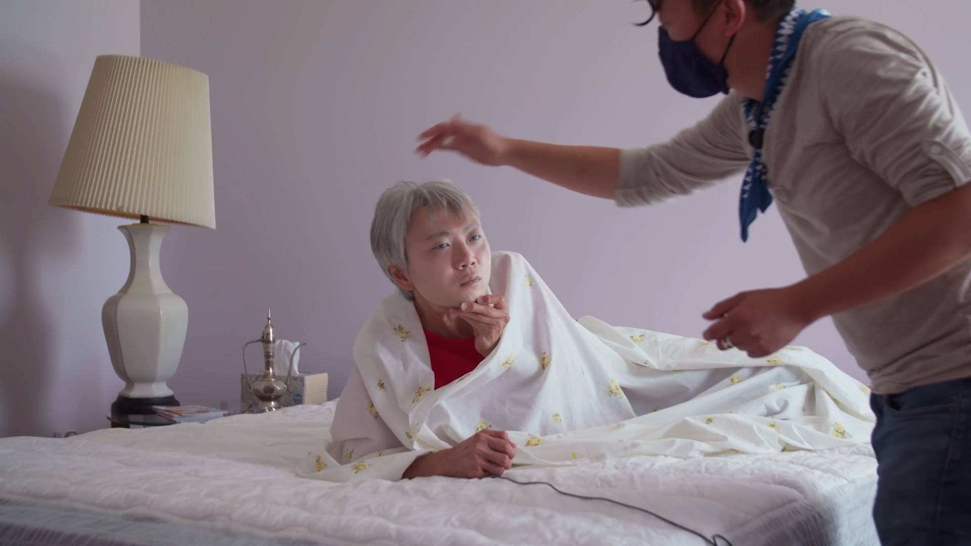 Tommy Kha setting up a scene to be photographed of model wrapped in covers on a bed holding a mask of the artist's face over their own.