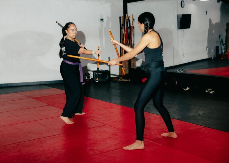 Two women practice martial arts with wooden sticks on red mats in a training studio, facing each other in a defensive stance.