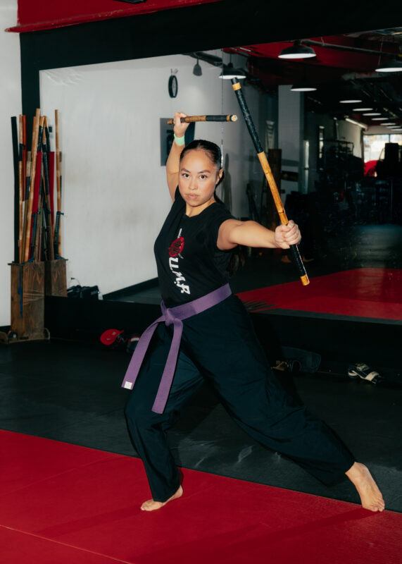 Caroline Garcia in martial arts attire with a purple belt holds two sticks in a defensive stance on a red mat in a training studio.