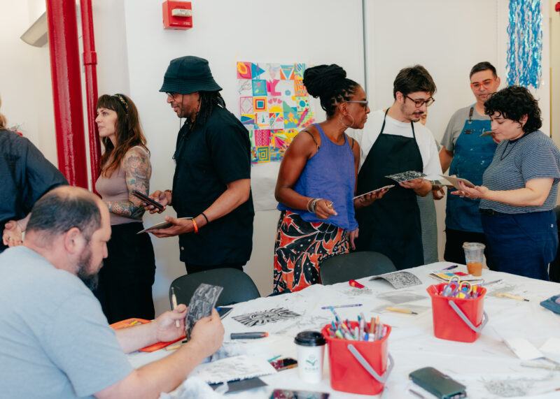 A group of adults stands and sits around a table with art supplies, examining and holding creative artworks in a bright room.