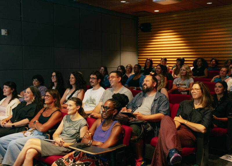 A group of adults sits in red theater seats, attentively watching a presentation in a modern lecture room.